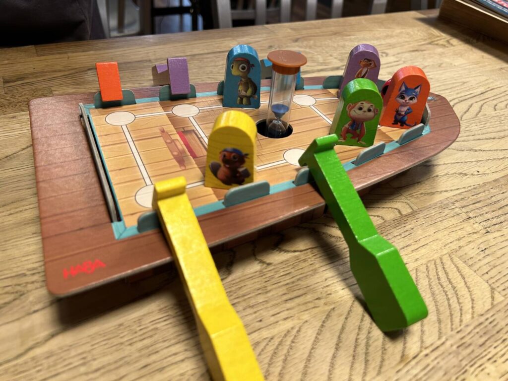 Chunky wooden tokens representing each of the crew members stand atop a tall, thick cardboard boat, with wooden paddles hanging off the sides. The paddles correspond in color to the tokens.