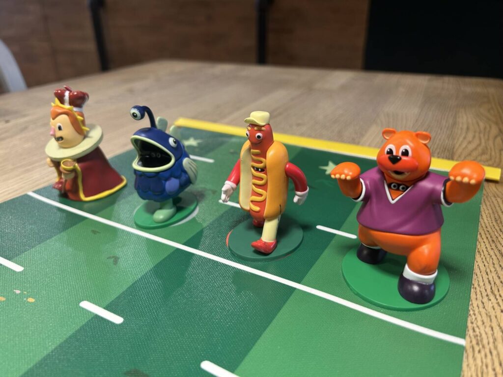 Four plastic figures, boldly colored, queue up at the starting line. The figures, from left to right, are a queen, an angler fish, a hot dog, and a bear.