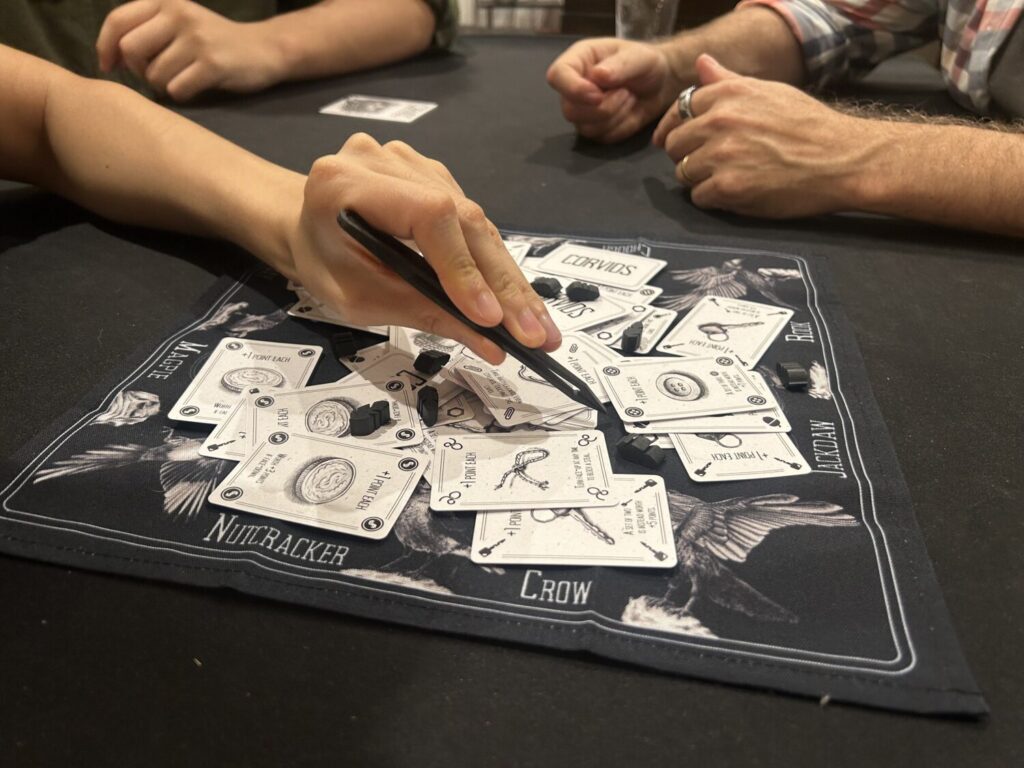 A hand holds long black tweezers, manipulating a card that sits among a large pile of cards on top of a black cloth matt.