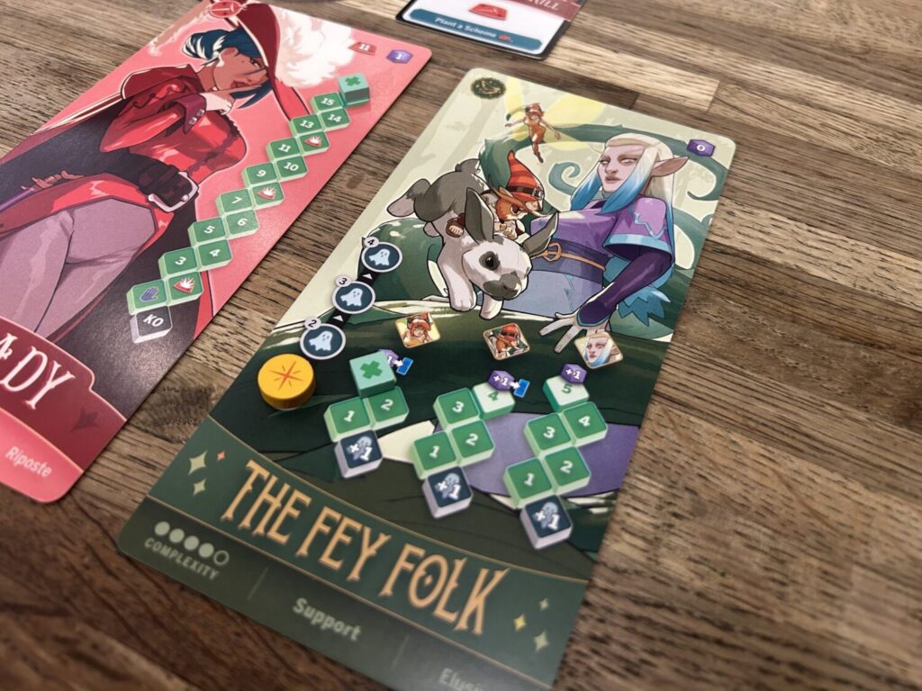 A pair of tall rectangular character boards lay next to each other on a wooden table. Each features a bold and colorful illustration and a health track.