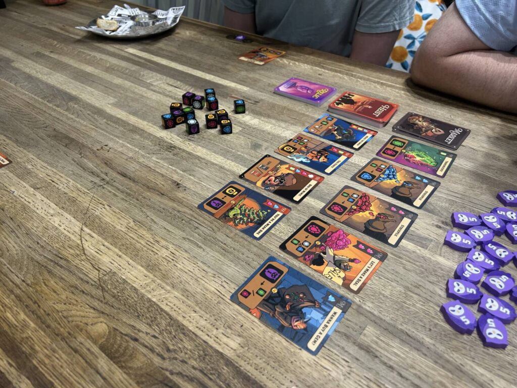 A table with two rows of cards, a large number of purple wooden owl tokens, and a large pile of dice.