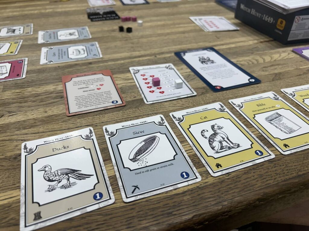 A number of cards on a wooden table. Some show items, some show health and reputation tracks.